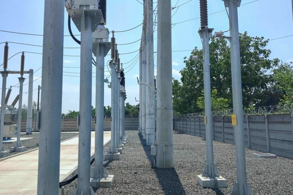 O serie de stâlpi metalici și piloni de beton susțin cabluri electrice într-o stație de transformare aflată pe un teren cu pietriș și un zid de beton.