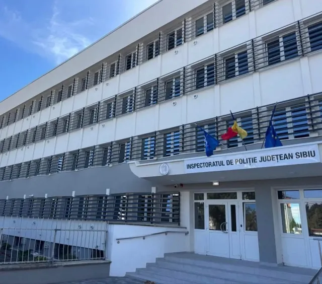 O clădire albă cu mai multe etaje prezintă ferestre cu jaluzele și trei steaguri suspendate deasupra unei intrări cu uși de sticlă și scări.