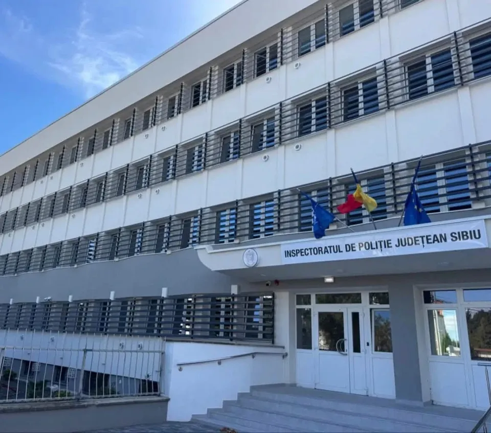 O clădire albă cu mai multe etaje prezintă ferestre cu jaluzele și trei steaguri suspendate deasupra unei intrări cu uși de sticlă și scări.