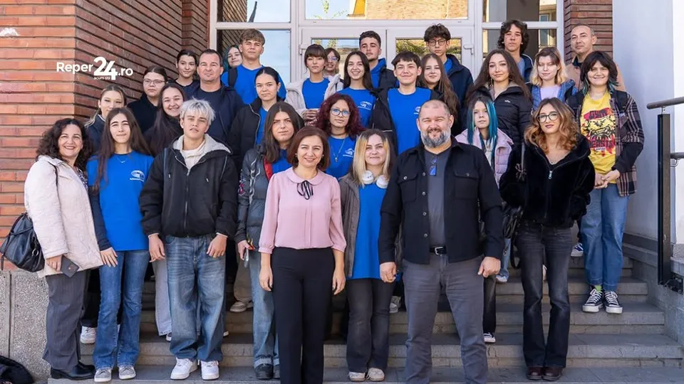Un grup de tineri stă în rânduri pe scări în fața unei clădiri cu ferestre mari și pereți din cărămidă.