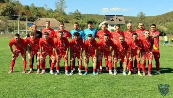 Un grup de fotbaliști în echipamente roșii și altele în albastru stau pe un teren de fotbal sub un cer senin.