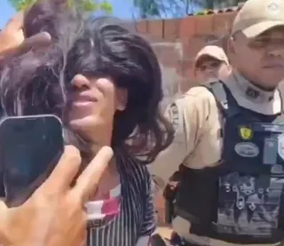 O persoană cu păr negru lung este fotografiată cu un telefon mobil, în timp ce un bărbat în uniformă și vestă tactică stă în spate.