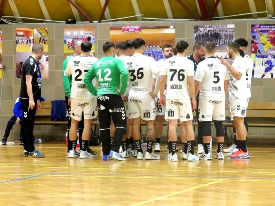Un grup de sportivi în echipamente albe și verzi stau întrupiți pe un teren de handbal în fața unui perete cu fotografii.