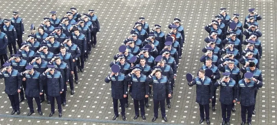 Grupuri de persoane îmbrăcate în uniforme închise la culoare stau în rânduri pe o suprafață pavată cu pietriș.
