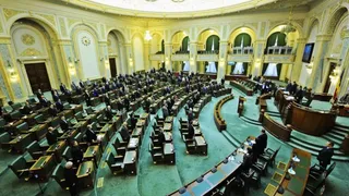 Imaginea de ansamblu cu o sală de ședință plină, cu persoane în costume formale așezate la bănci și la un pupitru mare.