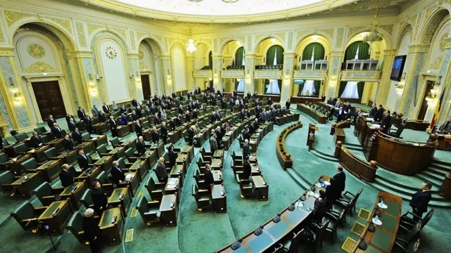 Imaginea de ansamblu cu o sală de ședință plină, cu persoane în costume formale așezate la bănci și la un pupitru mare.