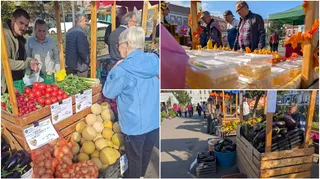 Oameni vizitează standuri de piață cu legume, fructe și produse diverse dispuse în cutii de lemn și pe mese în aer liber.