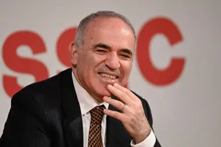 Un bărbat cu părul scurt și gri, purtând un sacou negru, cămașă albă și cravată brună, zâmbește cu mâna la nivelul bărbii.