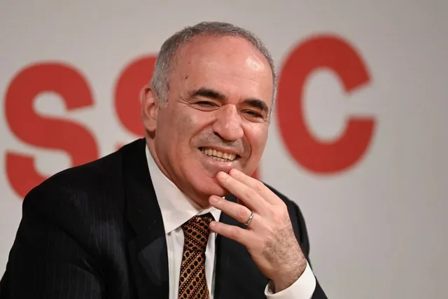 Un bărbat cu părul scurt și gri, purtând un sacou negru, cămașă albă și cravată brună, zâmbește cu mâna la nivelul bărbii.
