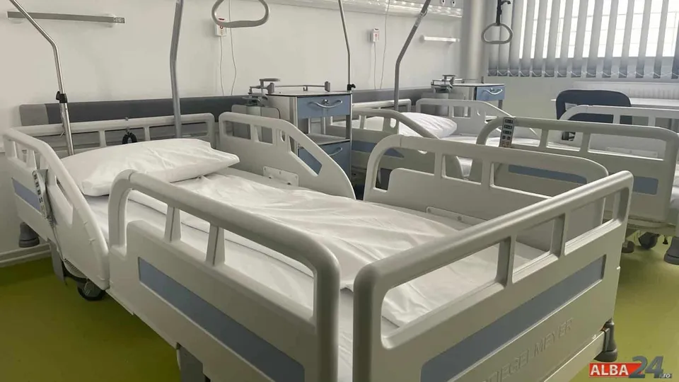 Paturi medicale albe, cu saltele acoperite, într-un salon cu ferestre și instalații montate pe perete.