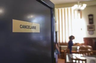 O placă cu textul CANCELARIE este atașată pe o ușă închisă, în timp ce o persoană stă în fundalul unei încăperi luminoase.