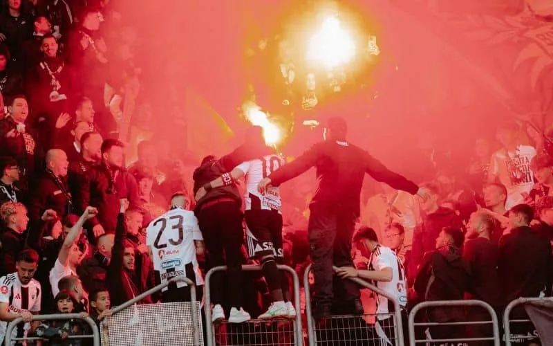 O mulțime de oameni stă în tribune sub o lumină roșie intensă, cu figuri siluete în prim-plan lângă bariere metalice.