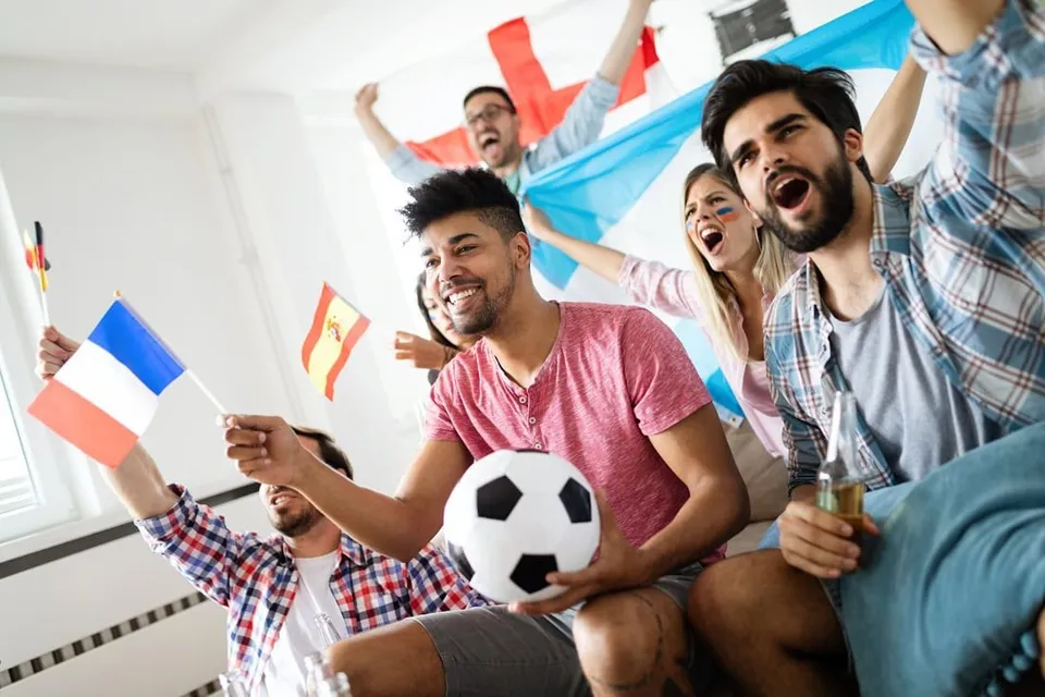 Un grup de tineri zâmbește și strigă, un bărbat ținând un fotbal alb cu pete negre, în timp ce alții ridică steaguri colorate.