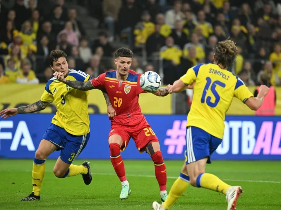 Trei jucători de fotbal dispută o minge pe un teren de gazon verde, fiind înconjurați de spectatori în tribunele din fundal.