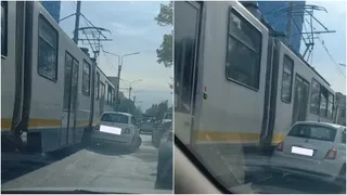 Un tramvai alb cu o dungă galbenă este poziționat lângă un autoturism alb pe o stradă cu vegetație și stâlpi de iluminat.