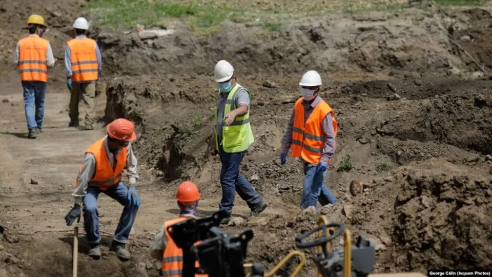 Cinci muncitori cu căști de protecție și veste reflectorizante lucrează pe un șantier cu pământ excavat.