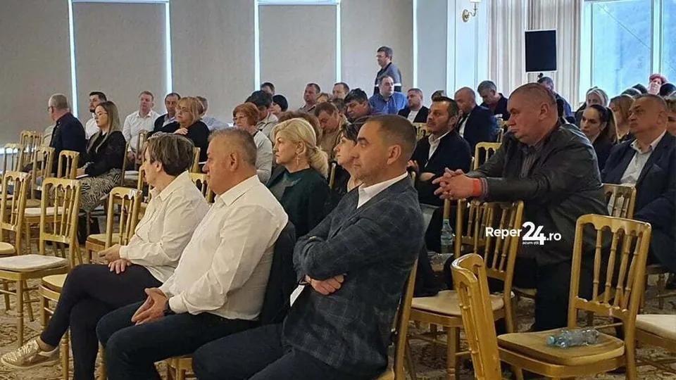Un grup de oameni stă așezat pe scaune de lemn într-o încăpere luminoasă, în timp ce un bărbat stă în picioare și vorbește.