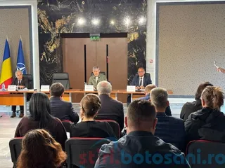 Un grup de persoane stă la mese lungi într-o sală de ședințe, în timp ce un public este așezat cu spatele la cameră.
