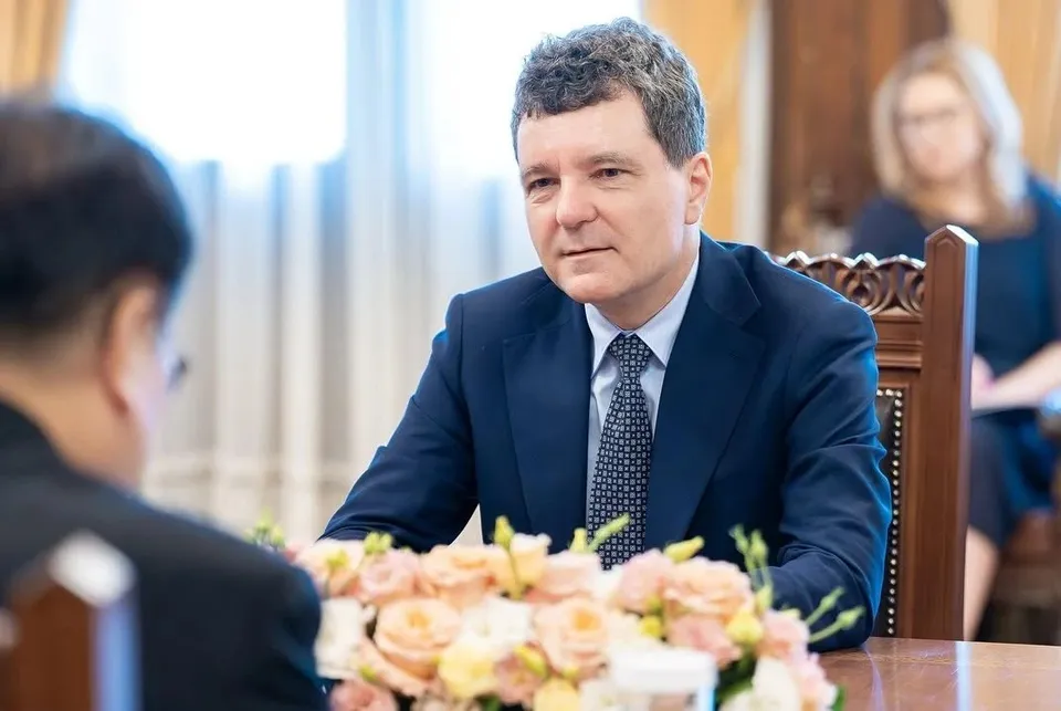 Un bărbat cu părul scurt și cămașă albă, purtând un sacou albastru și cravată cu model, stă la o masă lângă un buchet de flori.