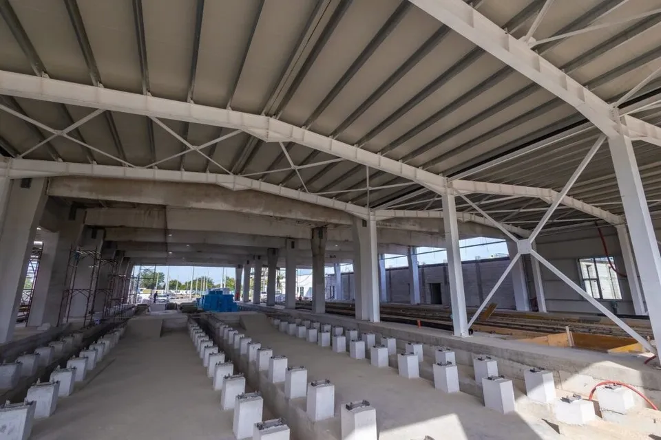 O structură metalică albă susține un acoperiș de plăci gri deasupra unei zone de construcție cu piloni de beton și elemente de fundație aliniate.