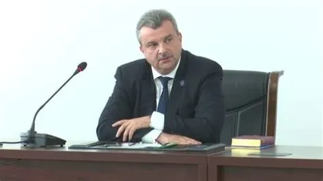 Un bărbat cu părul cărunt, purtând costum închis la culoare și cravată, stă la o masă de lemn lângă un microfon și un dosar.