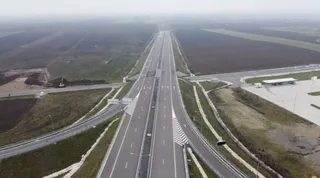 O vedere aeriană prezintă o autostradă cu mai multe benzi de rulare, delimitată de bariere metalice și înconjurată de câmpuri deschise sub un cer înnorat.