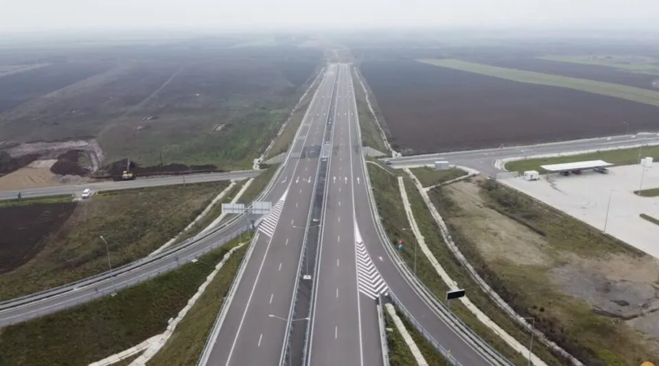 O vedere aeriană prezintă o autostradă cu mai multe benzi de rulare, delimitată de bariere metalice și înconjurată de câmpuri deschise sub un cer înnorat.