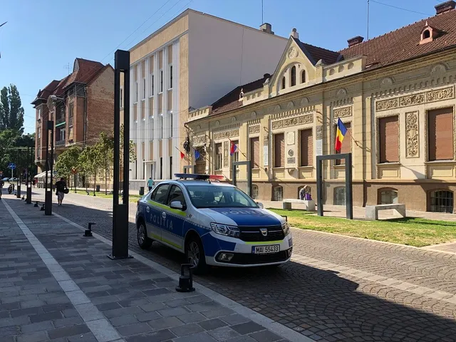O mașină de poliție albastră și albă este parcată pe un drum pavat în fața unor clădiri vechi și a unei construcții moderne sub un cer senin.