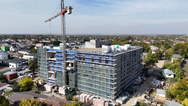 O clădire mare de beton cu schele metalice și o macara înaltă sunt situate într-o zonă rezidențială cu case mici și copaci.