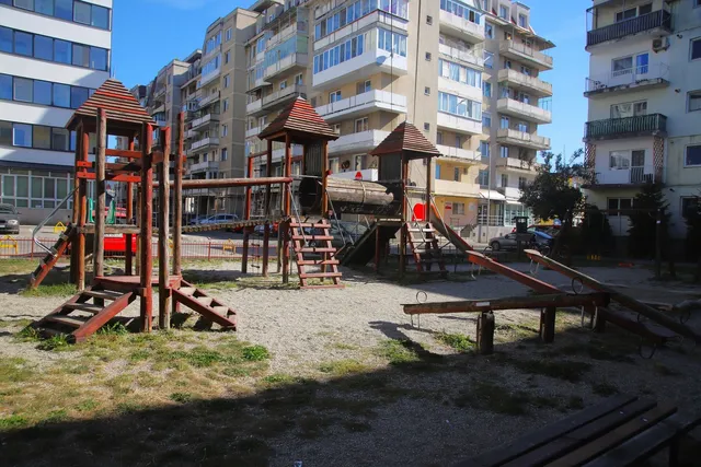 Un loc de joacă din lemn cu turnuri, scări și un balansoar se află într-un spațiu deschis lângă clădiri de apartamente înalte sub un cer senin.
