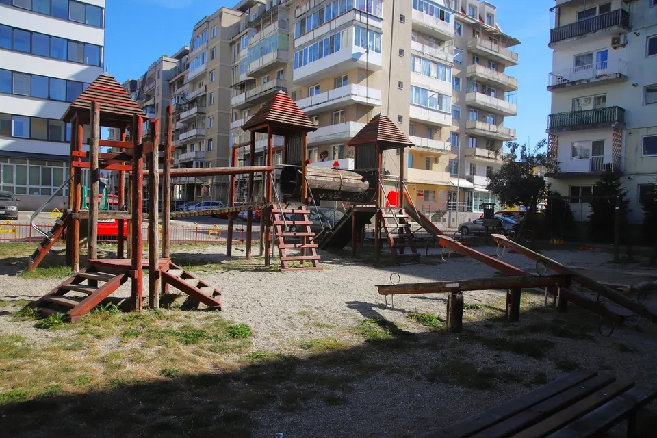 Un loc de joacă din lemn cu turnuri, scări și un balansoar se află într-un spațiu deschis lângă clădiri de apartamente înalte sub un cer senin.