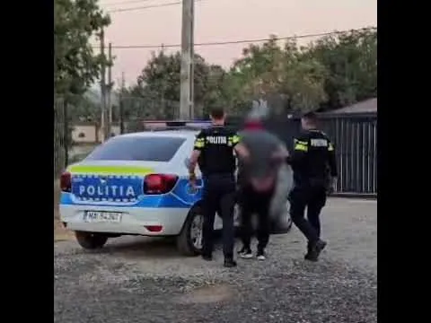 Unii polițiști în uniformă neagră cu inscripții galbene îl conduc pe un bărbat într-o poziție ușor aplecată lângă o mașină de poliție albă și albastră.