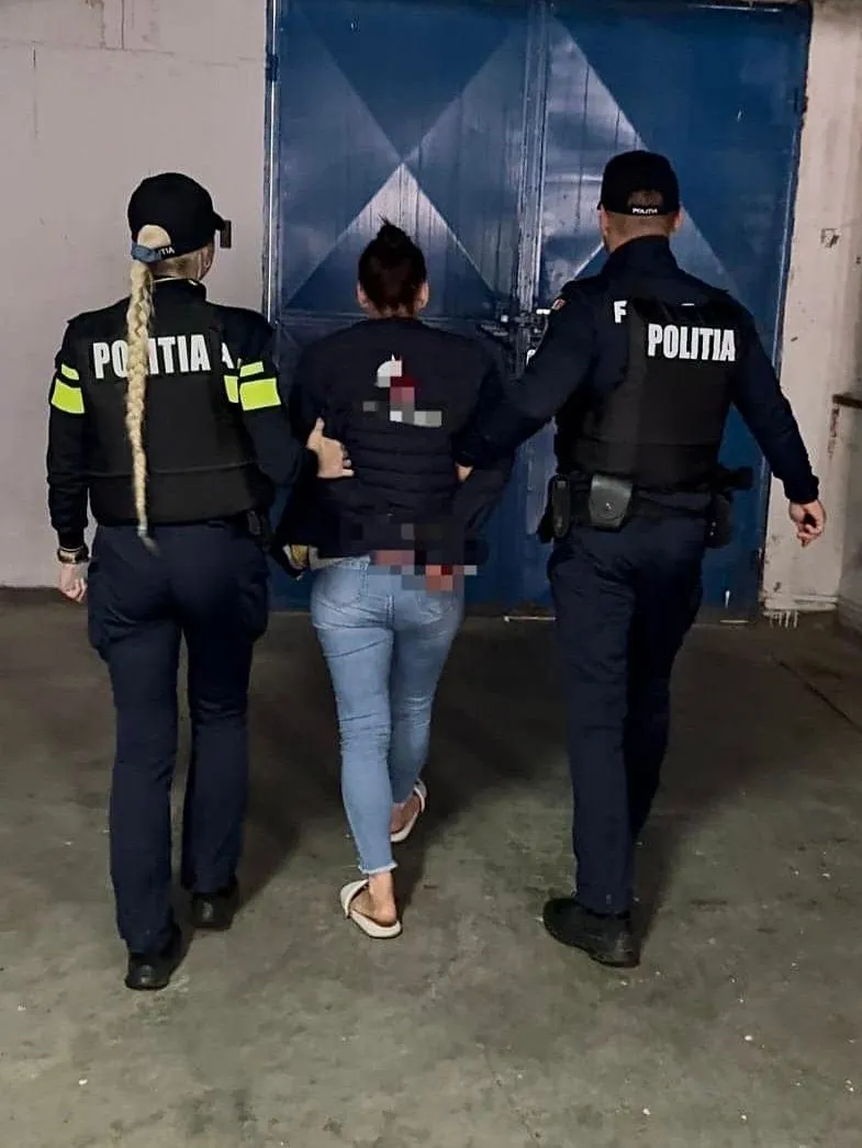 Trei persoane, două în uniformă de poliție și una în blugi și tricou negru, merg pe un drum spre o poartă albastră.