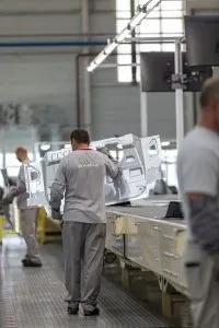 Un bărbat cu haine de lucru gri manipulează o piesă metalică de caroserie într-o fabrică, lângă alți muncitori.