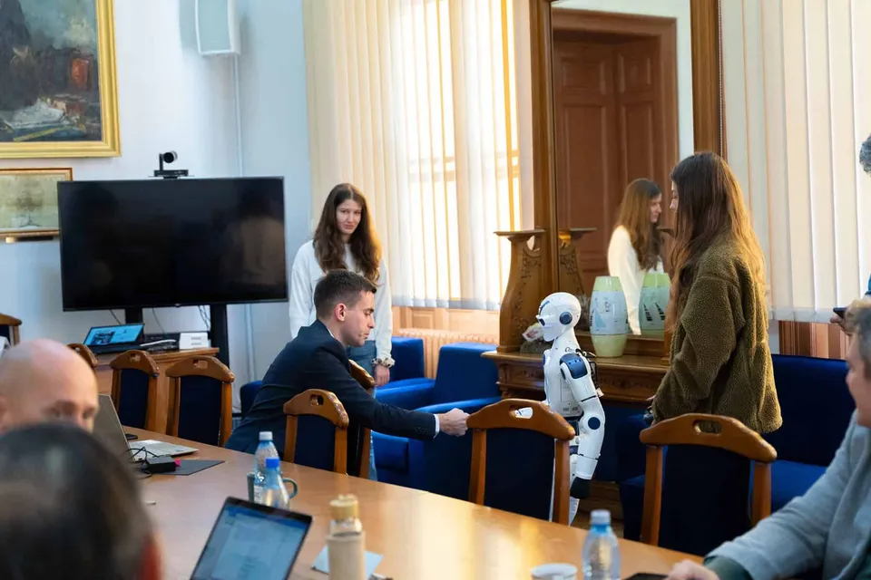 Un robot humanoid alb stă lângă o masă de lemn în presence de trei femei și un bărbat într-o încăpere cu ferestre mari.