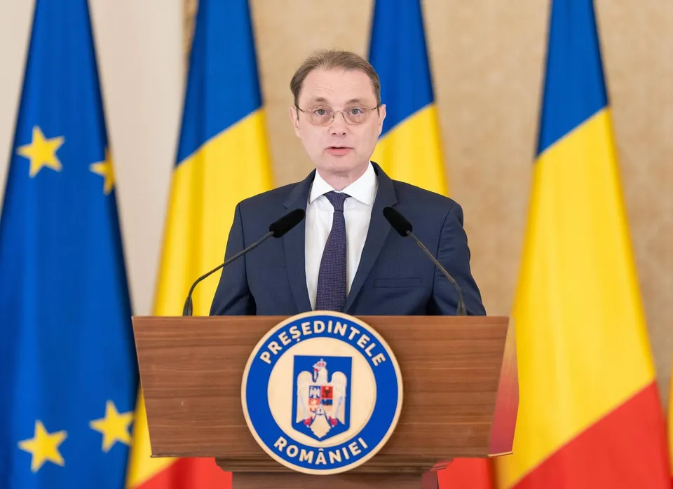 Un bărbat cu ochelari și cravată purtând un costum în fața unor steaguri, stând în spatele unui podium cu un emblema circulară.