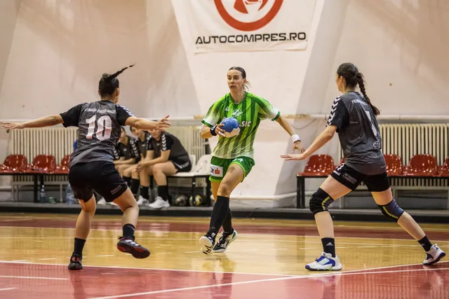 Trei jucătoare de handbal sunt pe terenul de sport, una cu o minge albastră în mână, fiind încadrate de alte două jucătoare.