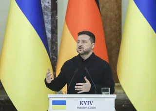 Un bărbat cu barbă și haină neagră stă în spatele unui podium alb lângă steaguri galben și albastru.