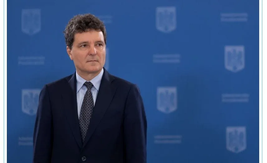 Un bărbat cu părul șaten și scurt, purtând un costum închis la culoare și o cămașă cu cravată, stă în fața unui fundal albastru cu embleme.