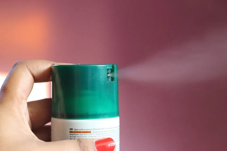 O mână ține un flacon mic de culoare verde și albă din care se degajă un spray fin într-un fundal roz.