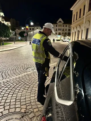Un agent de poliție cu vestă reflectorizantă verifică un vehicul negru pe un drum pavat noaptea.