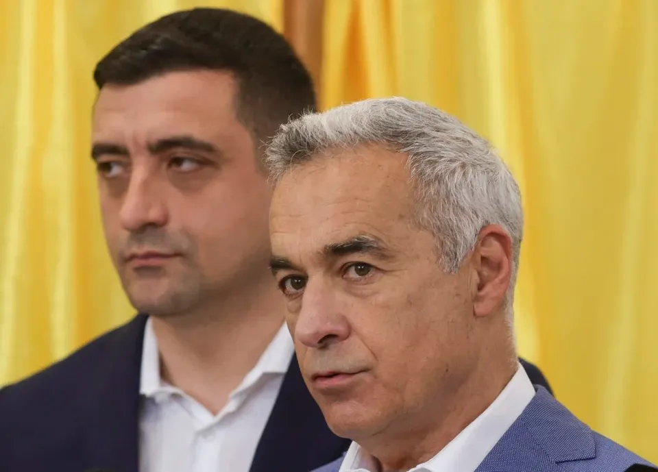 Un bărbat cu părul cărunt și o cămașă albă este prezentat în prim-plan, lângă un bărbat cu părul scurt și sacou închis la culoare.