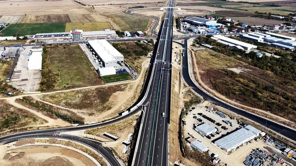 O vedere aeriană prezintă o autostradă cu mai multe benzi care traversează un peisaj cu terenuri agricole, clădiri industriale și drumuri de bağlantă.