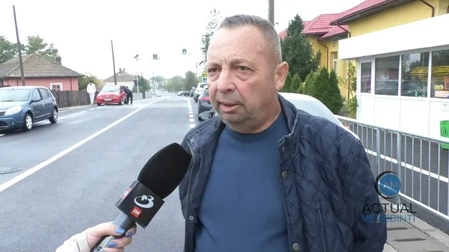 Un bărbat cu o jachetă albastră și un tricou închis la culoare este intervievat cu un microfon în fața unei străzi cu mașini parate.