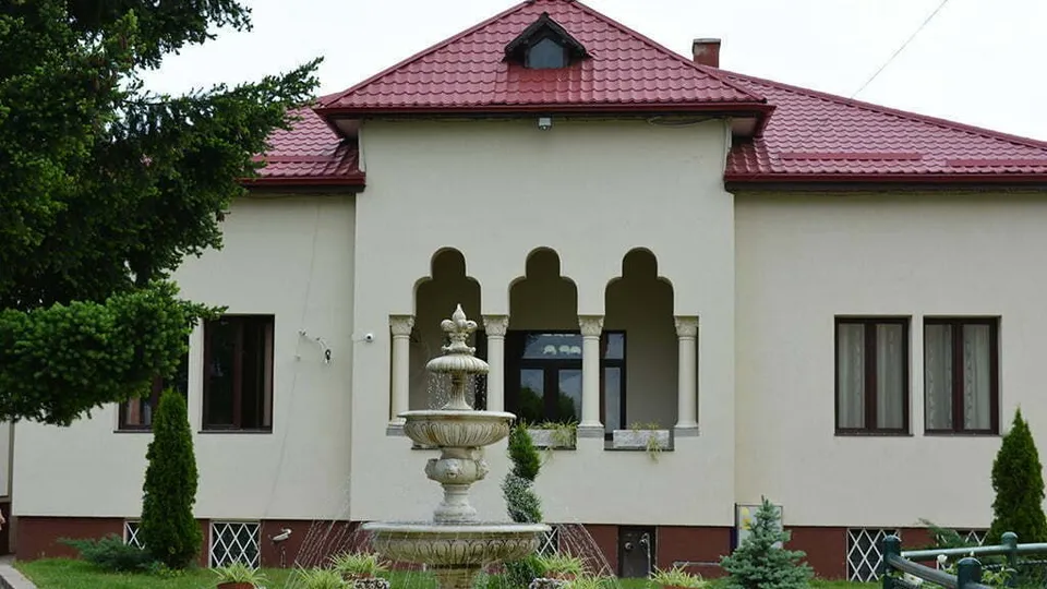 O clădire cu acoperiș roșu și un fântâi de piatră situat în fața acesteia, înconjurat de vegetație și un gard mic.