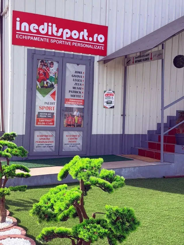 O clădire cu un panou roșu pe care scrie ineditsport.ro și o ușă de sticlă cu diverse reclame și imagini de sportivi.
