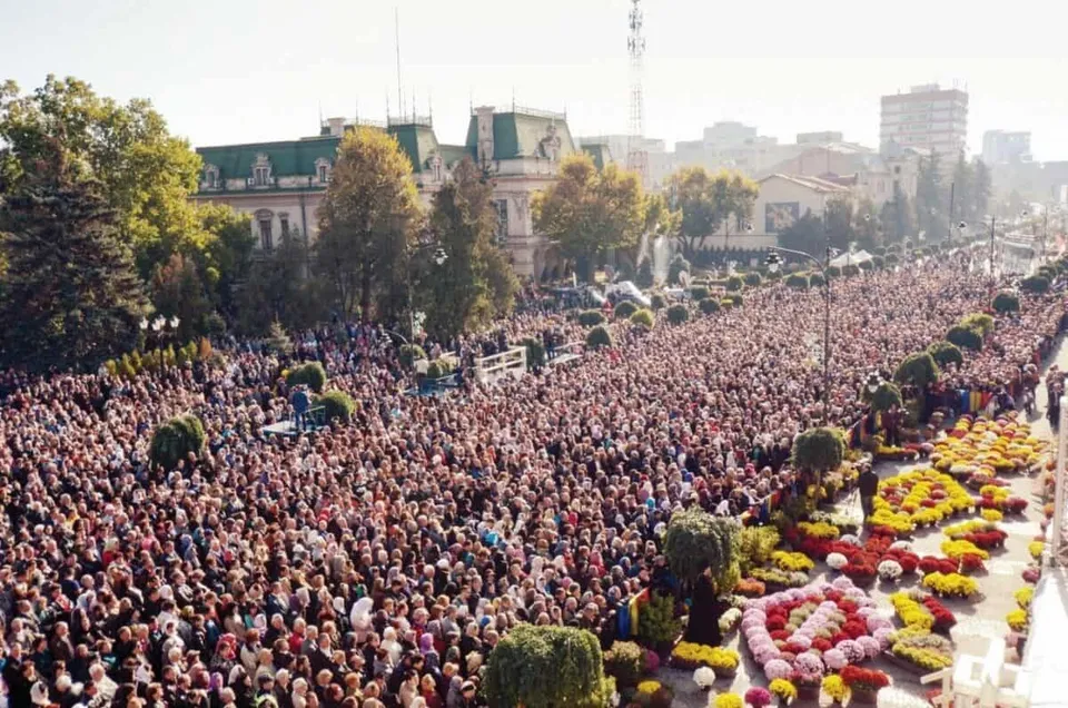 O mulțime mare de oameni este adunată pe o stradă lată lângă straturi de flori colorate și copaci decorați.