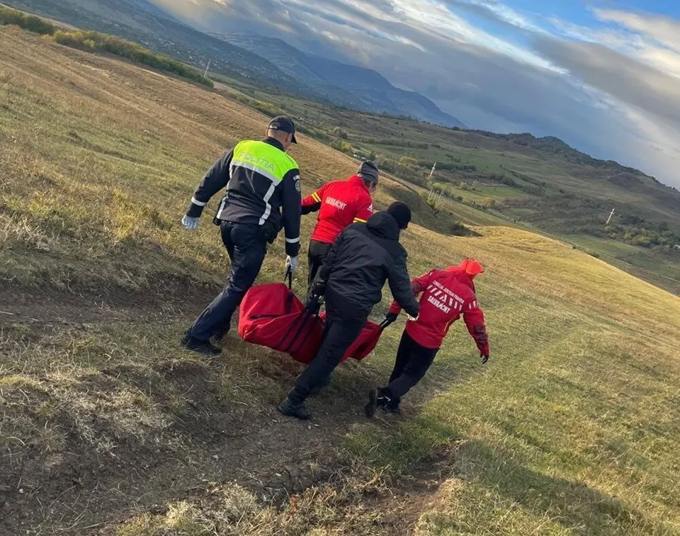 Patru persoane purtând haine de lucru colorate transportă un sac de transport roșu pe un teren înclinat acoperit cu iarbă uscată.