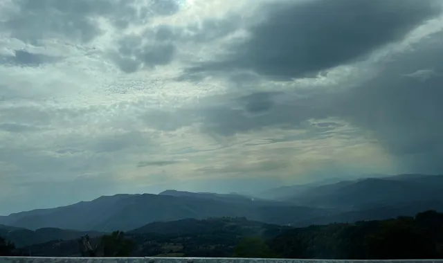 Un cer acoperit de nori gri și albi deasupra unui peisaj format din dealuri și munți întinși sub o lumină difuză.