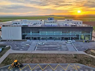 O clădire modernă de aeroport cu fațadă de sticlă este situată în fața unui teren de asfalat sub un cer la apus.